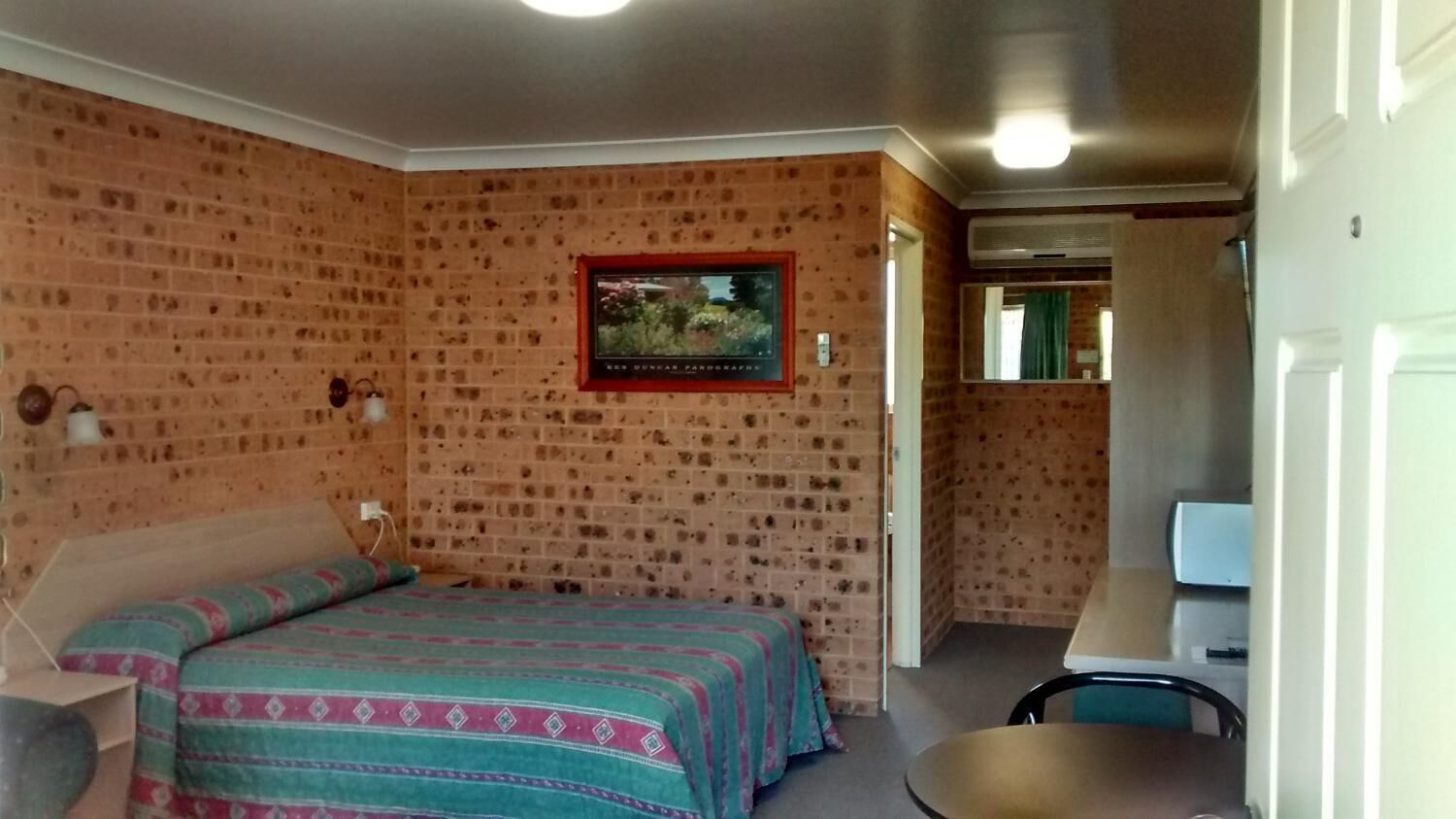 A room with a brick-patterned wall, a bed with a patterned bedspread, a small table, and a framed picture on the wall.