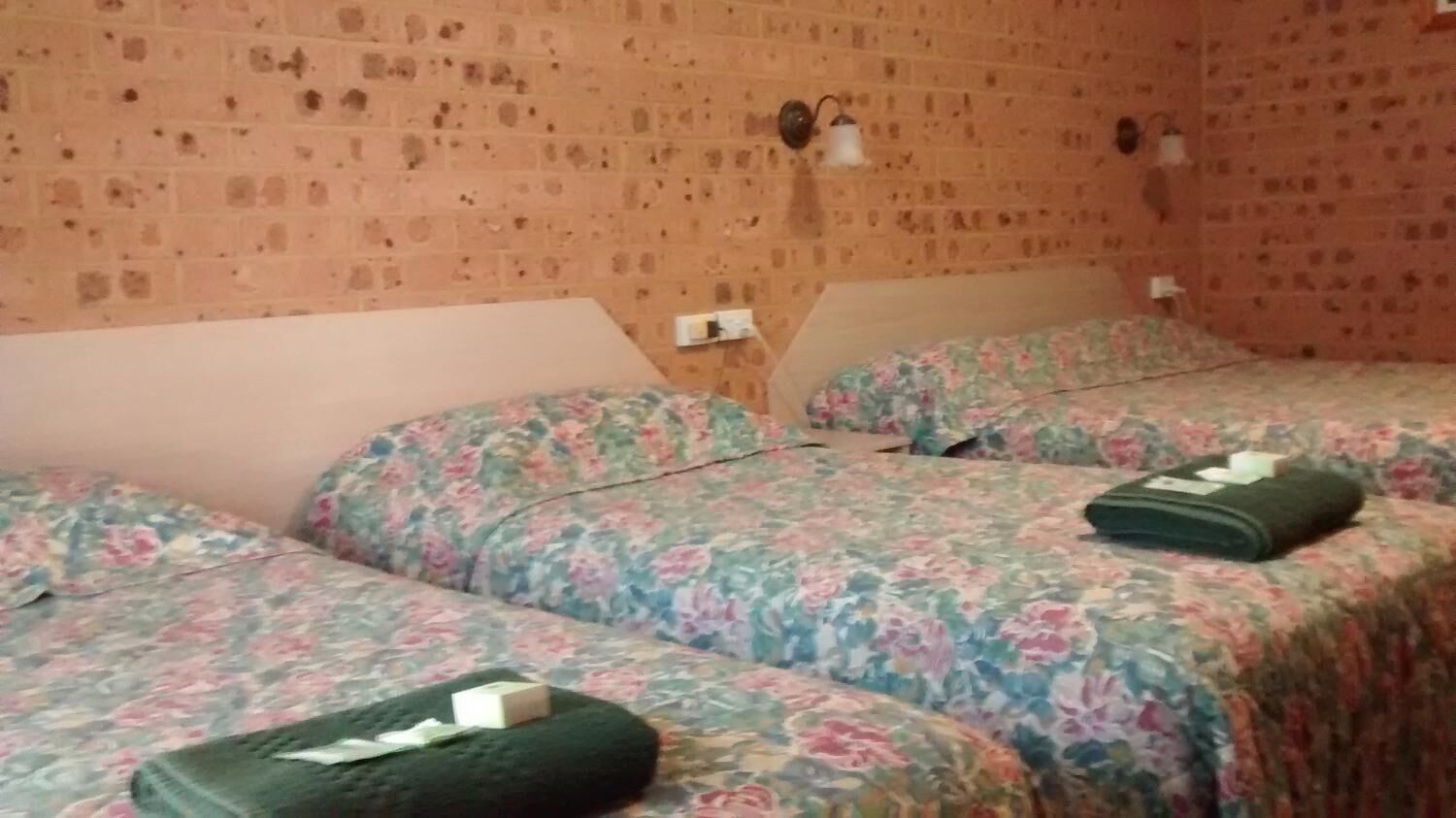 A room with three beds, each with floral bedding and a green pillow. The walls are brick-patterned, and there are wall-mounted light fixtures.