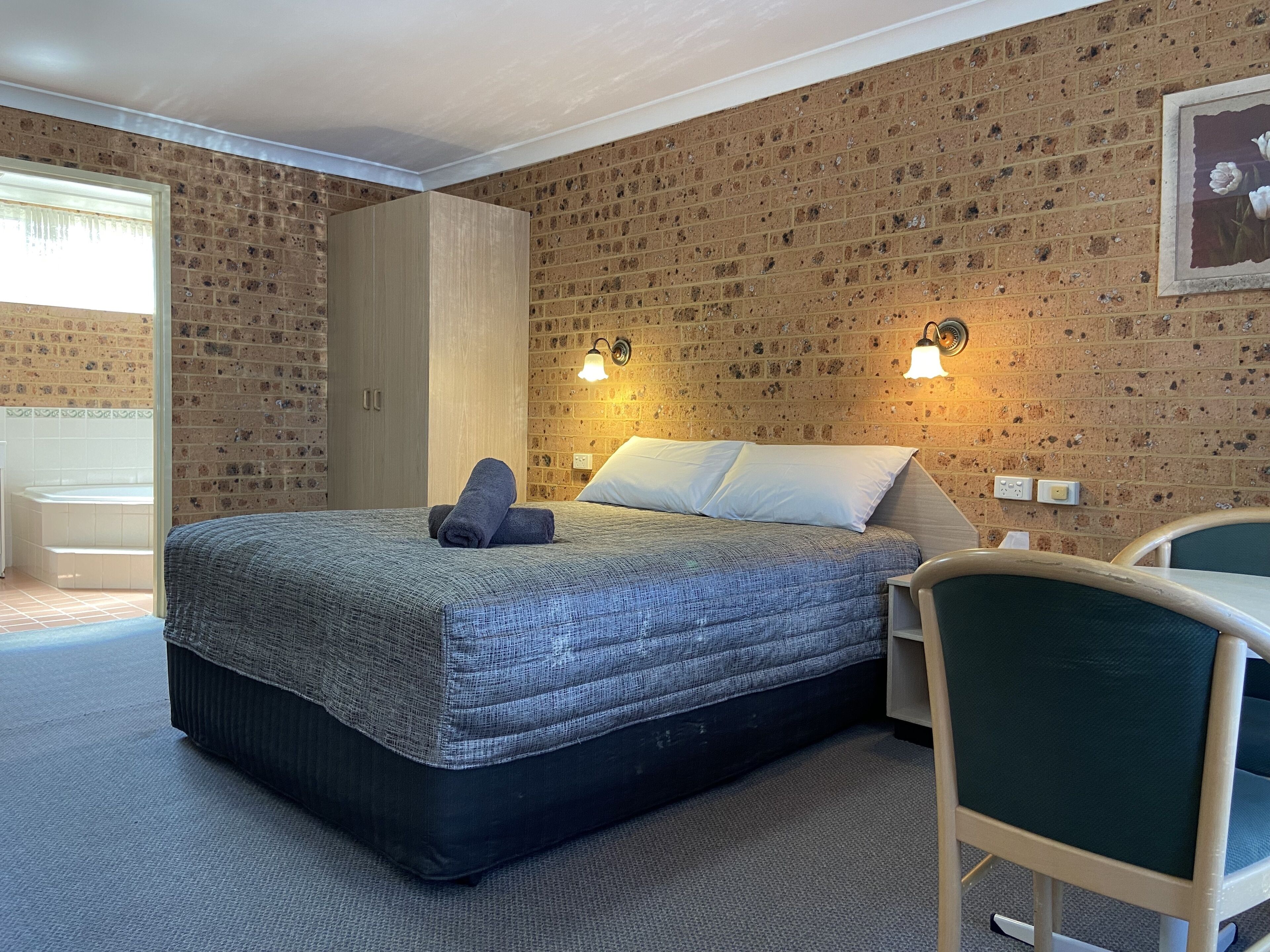 Un dormitorio con pared de ladrillo visto, una cama con colcha gris, dos almohadas, un armario de madera y una mesita de noche con lámpara.