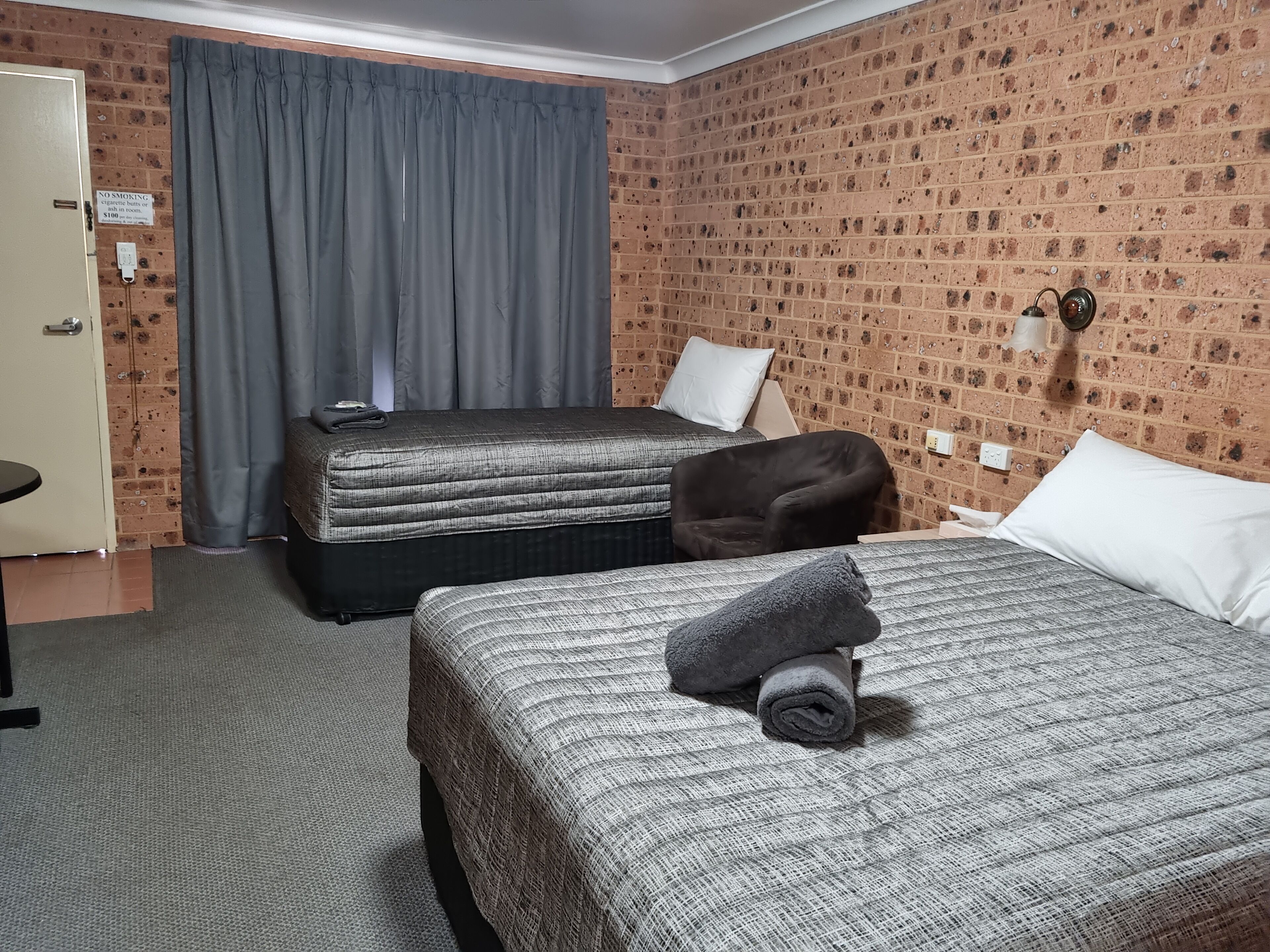 A hotel room with two beds, a grey curtain, a brick-patterned wall, and a no-smoking sign.