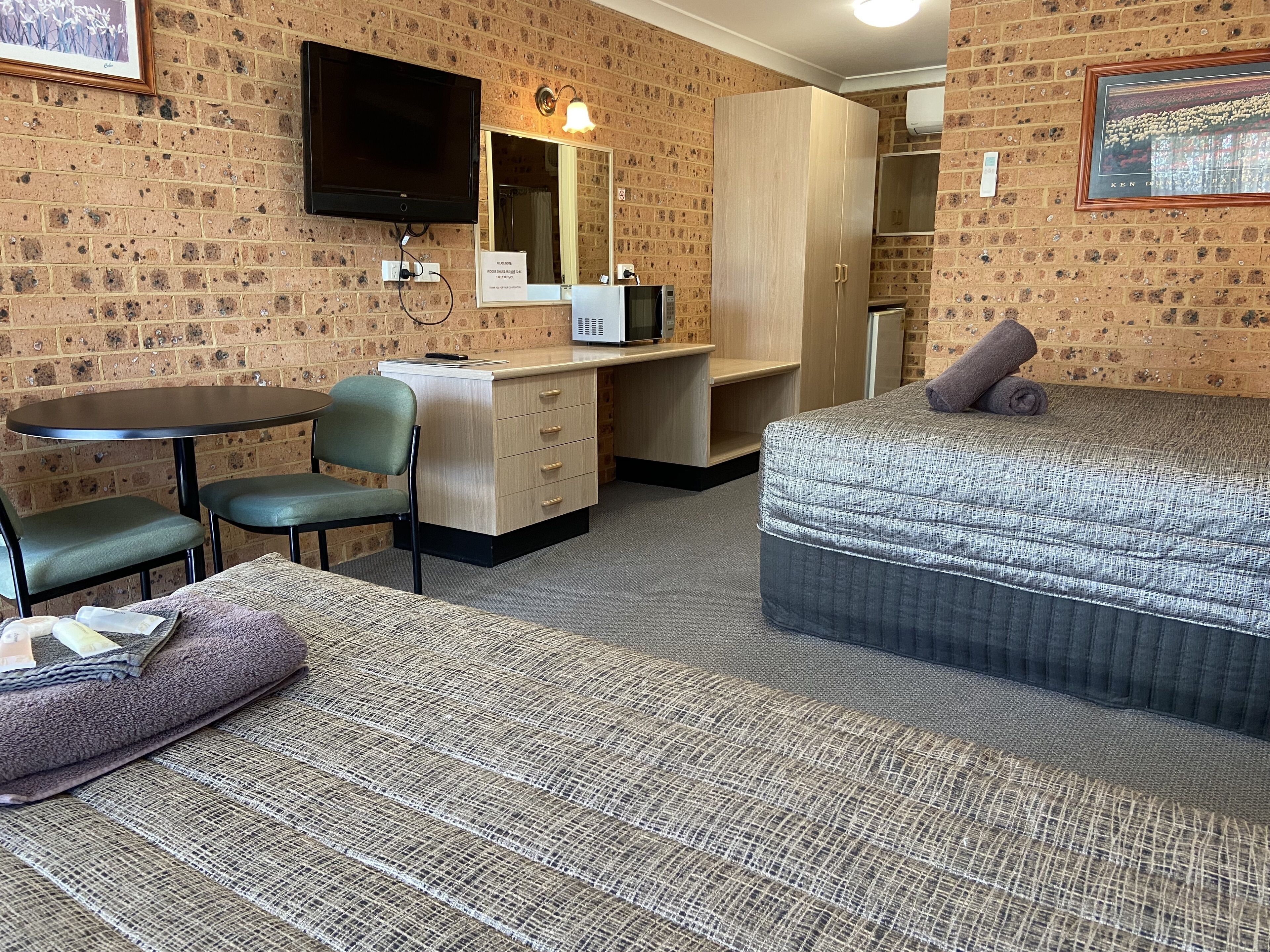 Habitación de hotel compacta con pared de ladrillos, televisor de pantalla plana, escritorio con silla, cama con almohada y pequeña mesa con silla.