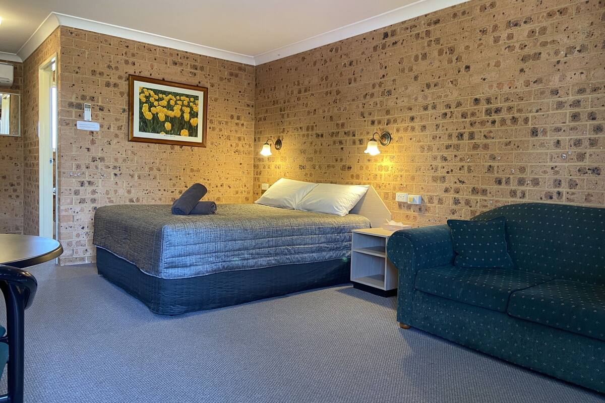 A room with a brick-patterned wall, a bed with a grey quilt, a green sofa, and a framed picture of flowers.