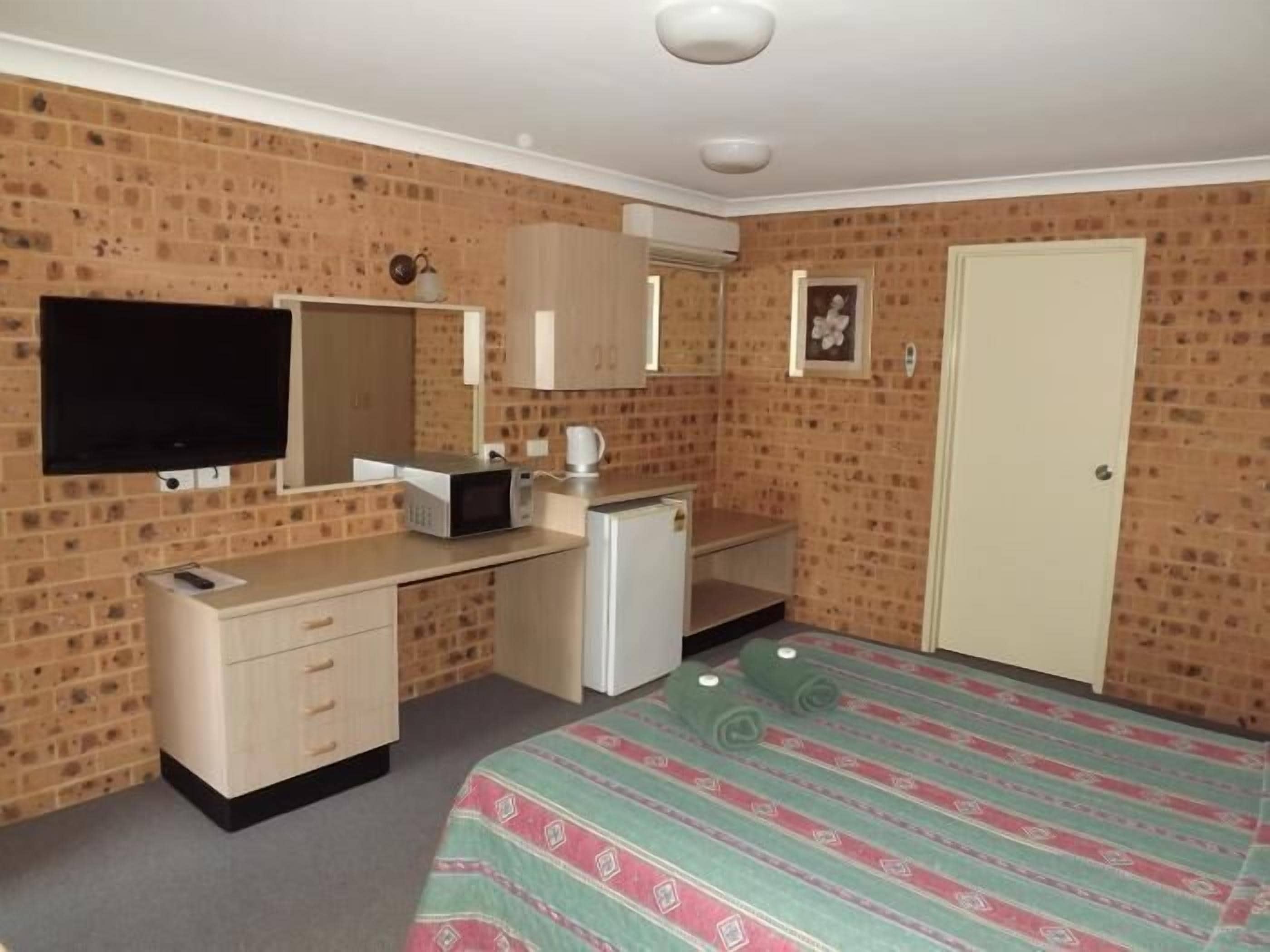 Un dormitorio con pared de ladrillo, una cama con colcha estampada, un escritorio pequeño con microondas, televisor y una puerta.