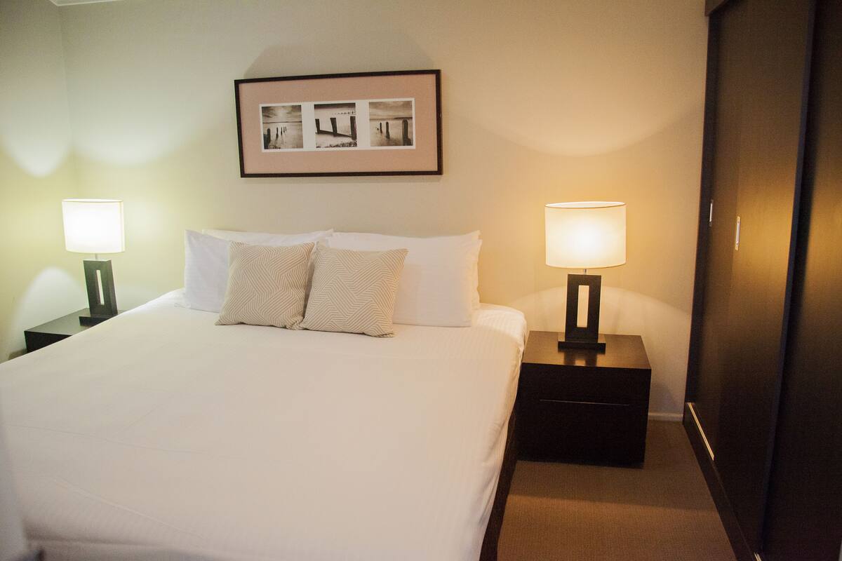 Ein ordentlich gemachtes Bett mit weißen Leinen und gemusterten Kissen, flankiert von zwei Nachttischen mit Lampen, in einem Hotelzimmer mit einem gerahmten Bild an der Wand.