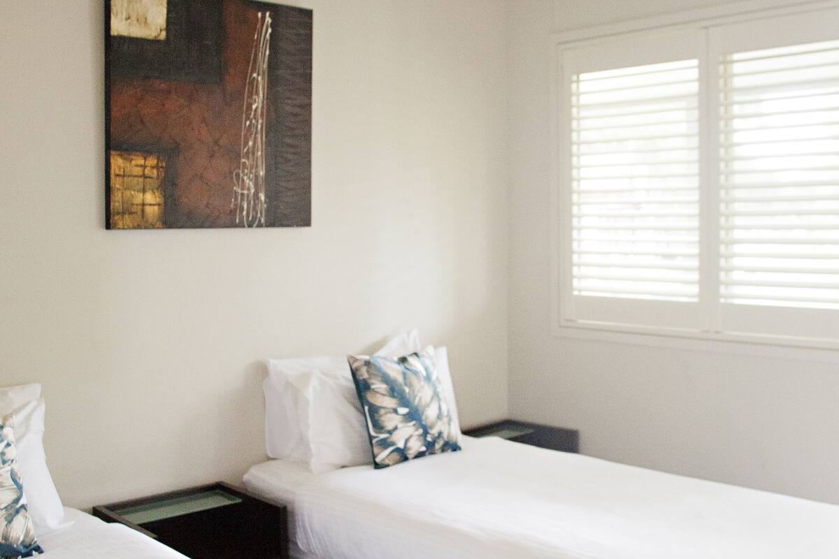 Ein Hotelzimmer mit zwei Einzelbetten, einem Deckenventilator, einem Fenster mit Jalousien und einem Bild an der Wand.