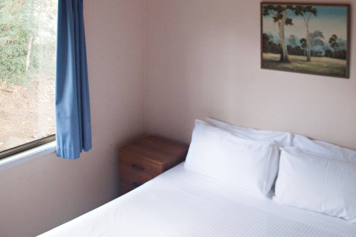 Ein Schlafzimmer mit einem Bett, einem Fenster mit blauer Vorhang, einem kleinen Nachttisch aus Holz und einem Baum-Gemälde an der Wand.