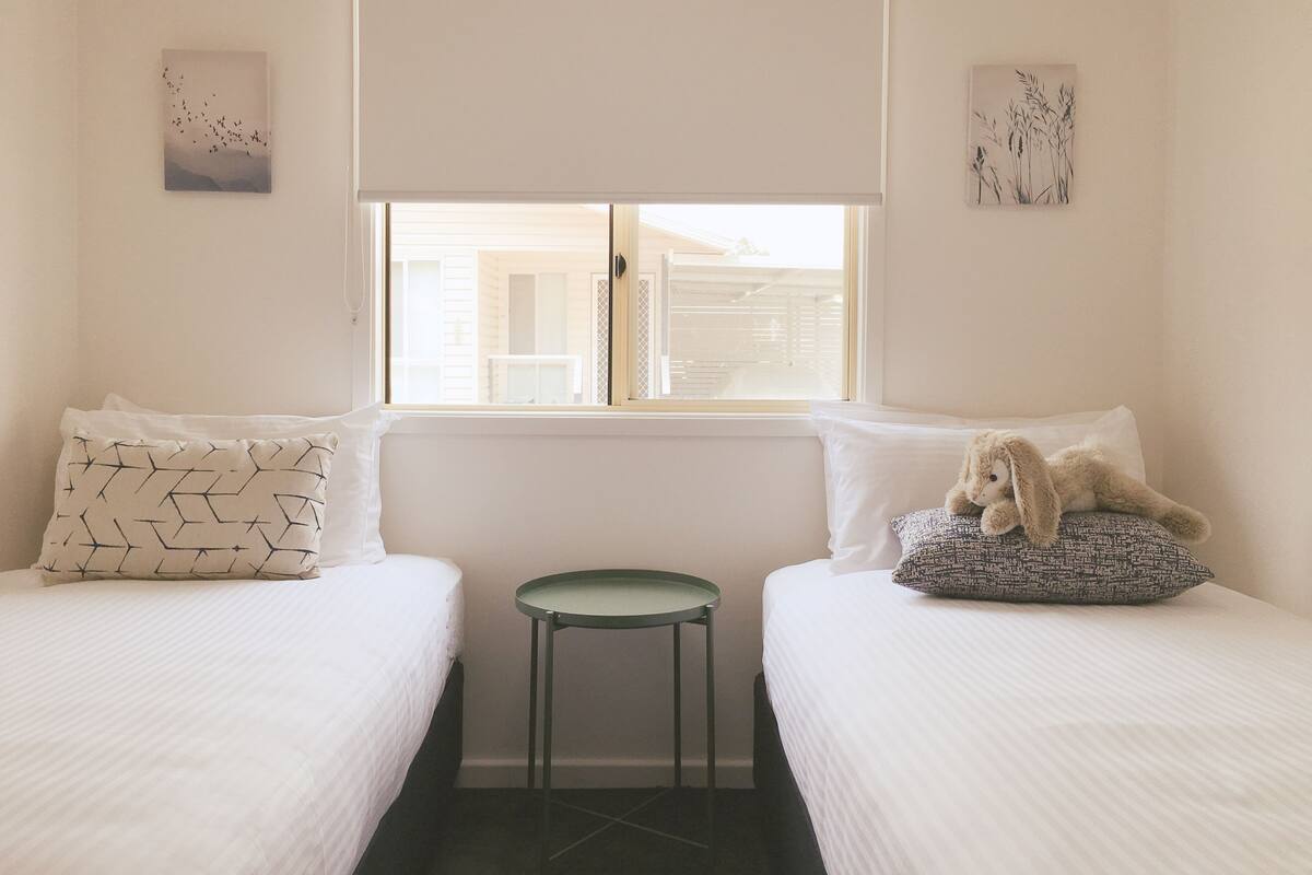 Ein Schlafzimmer mit zwei Betten, einem kleinen runden Tisch und einem Fenster mit Blick auf ein Gebäude.