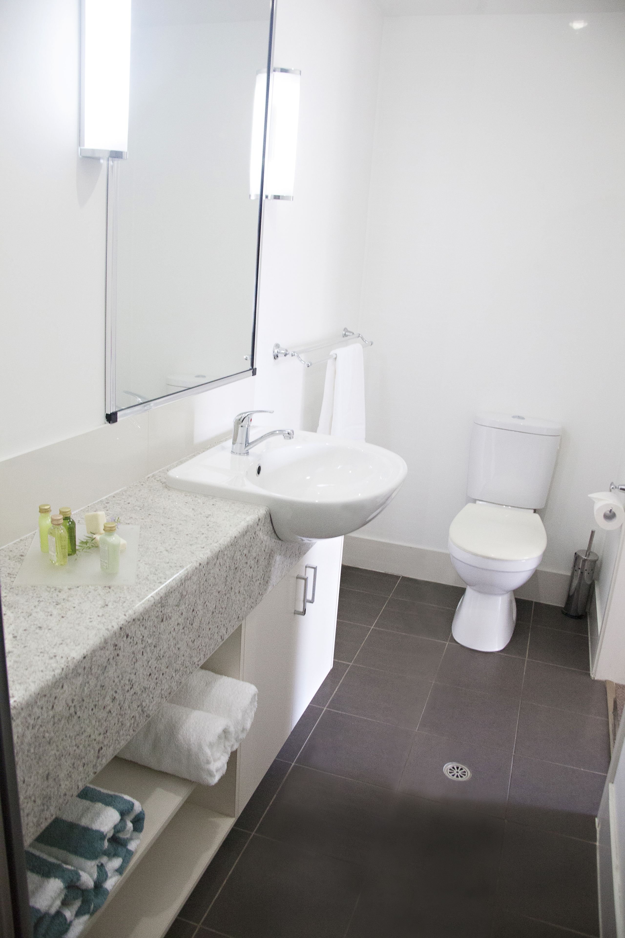 Uma casa de banho moderna com bancada de granito, lavatório branco, espelho, sanita branca e suporte para toalhas.