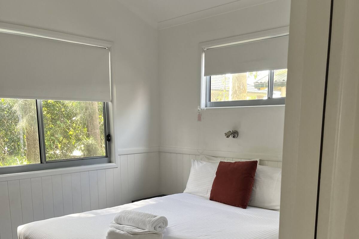 Ein Schlafzimmer mit einem großen Bett, weißer Bettwäsche, einem roten Kopfkissen und einem Fenster mit Blick auf Bäume.