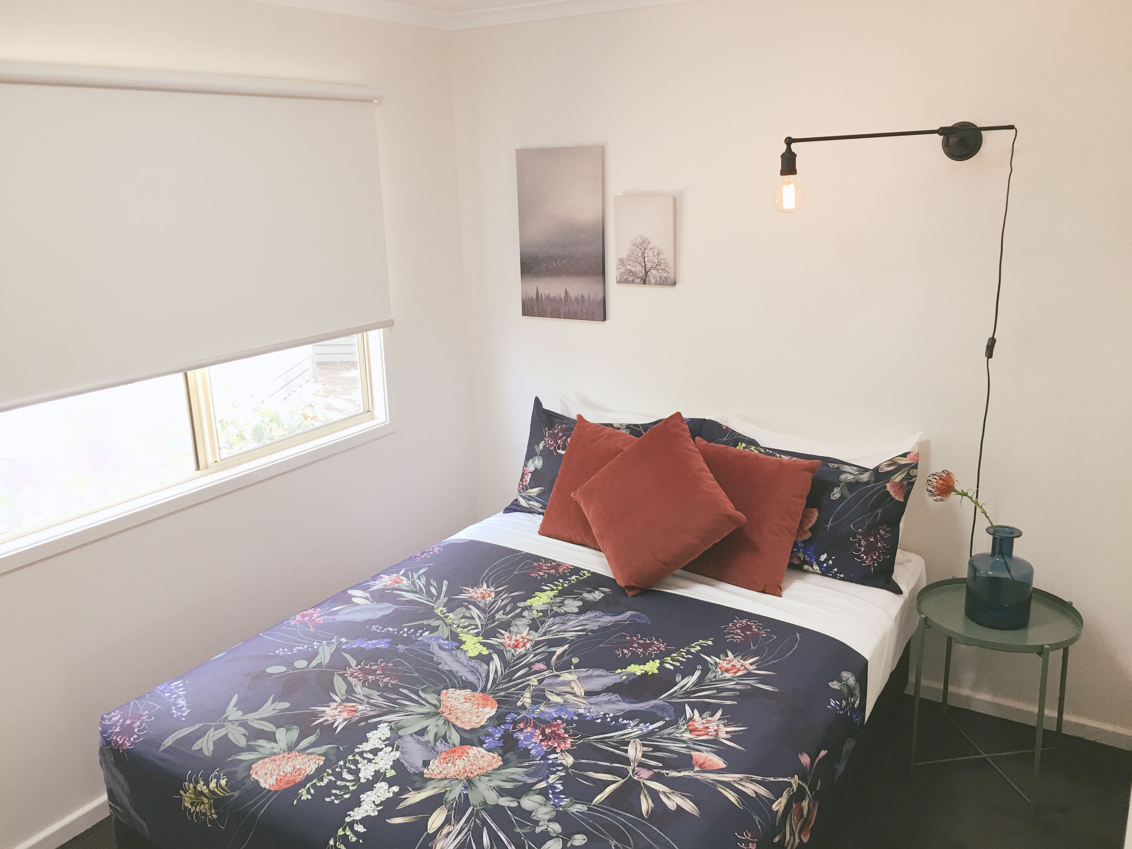A bedroom with a bed, a window, a wall-mounted lamp, and a small table with a vase.