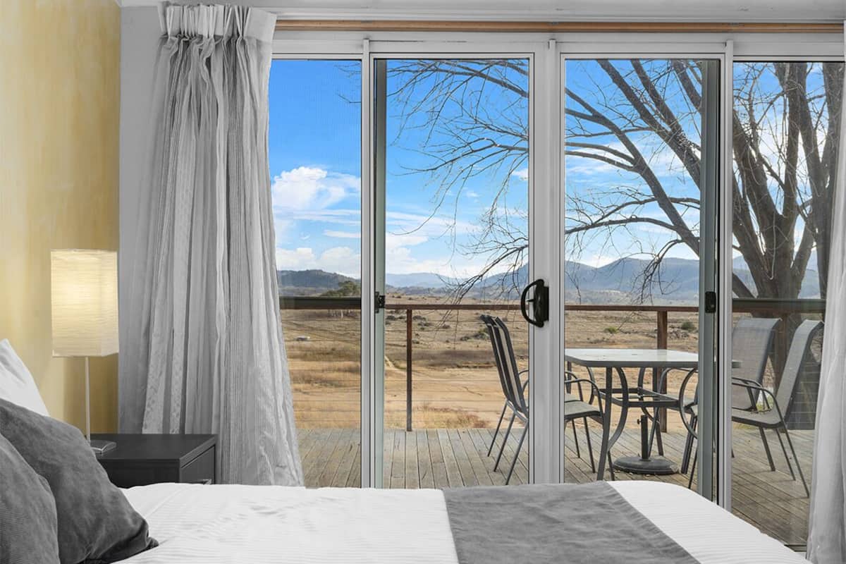 A bedroom with a large bed, a nightstand, a lamp, and a view of a deck with chairs and a table.