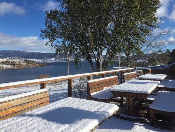 A snow-covered outdoor seating area with wooden benches and tables, overlooking a body of water and distant mountains. - Siesta Villa (East Jindabyne)