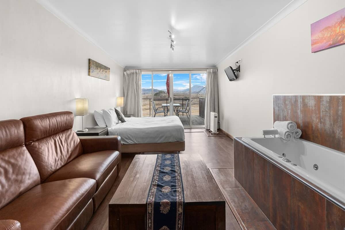 A modern hotel room with a leather sofa, a bed with a view, a wooden coffee table, a bathtub, and a dining area with a table and chairs.