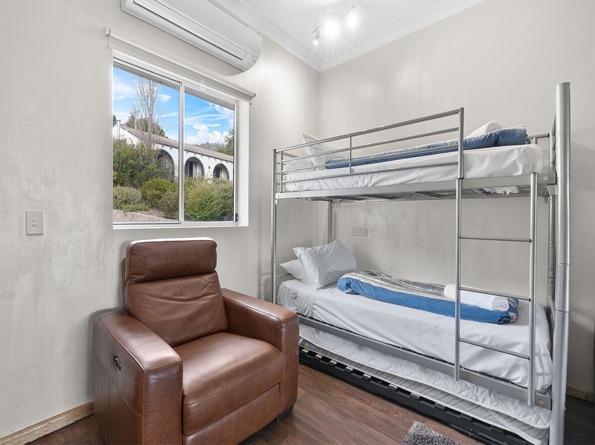 A bunk bed with white bedding, a brown leather chair, and a window with a view of greenery and a building.