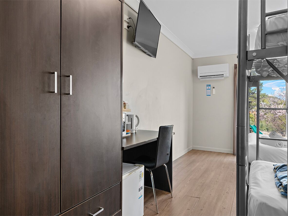 A compact room with a bed, a desk, a chair, a TV, and an air conditioning unit.