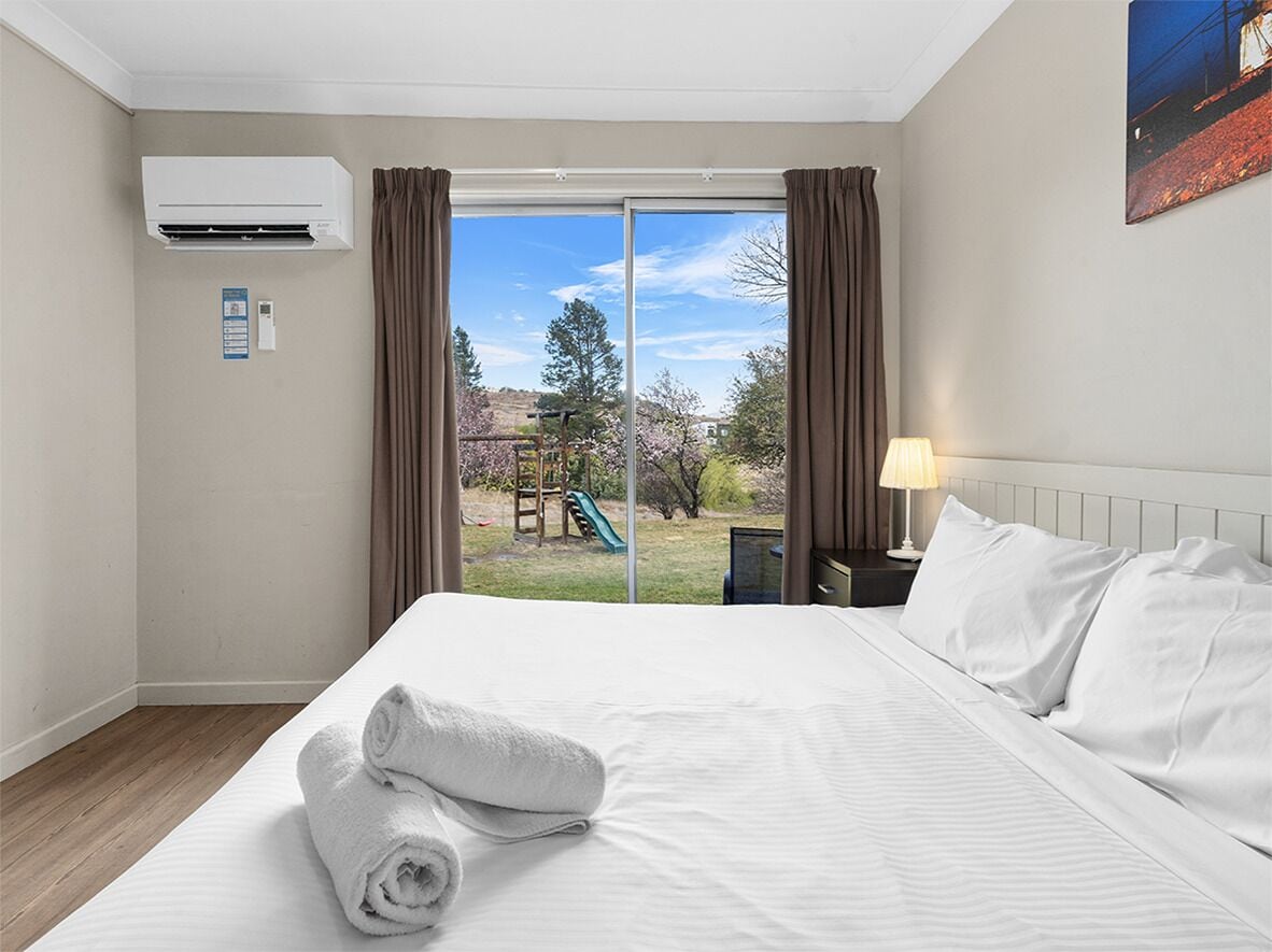 A hotel room with a large bed, a view of a playground, and an air conditioning unit.