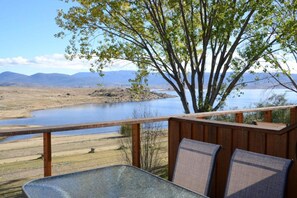 Una terraza de madera con una mesa de vidrio y dos sillas, con vistas a un lago y montañas distantes.