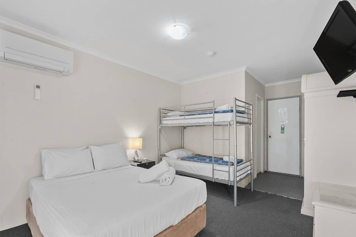 A hotel room with a single bed, a bunk bed, a desk with a lamp, a television, and an air conditioning unit.