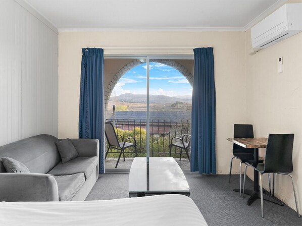 A hotel room with a bed, a sofa, a table, and chairs. There is a large window with curtains, and an air conditioning unit on the wall. - Siesta Villa (East Jindabyne)