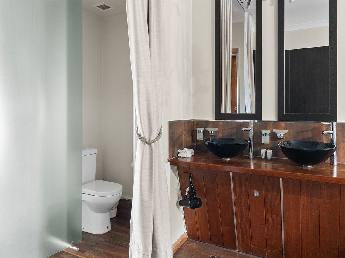 Un baño con tocador de madera, dos lavamanos tipo cuenco negros, un espejo y un inodoro blanco.
