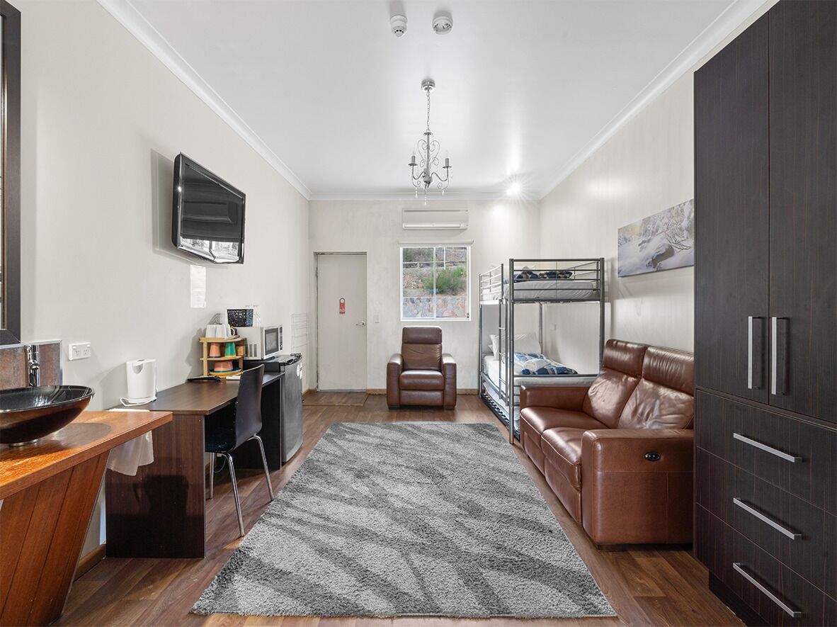 Ein kompakter Wohnbereich mit Küchenzeile, einer Sitzecke mit Sofa und Sessel, einem Etagenbett und einem dunklen Holzschrank.