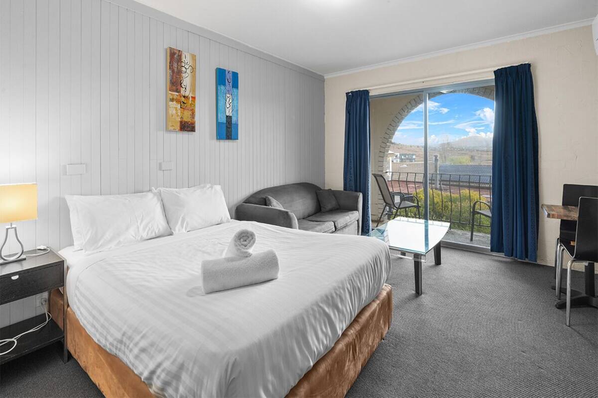 A hotel room with a bed, a desk, a chair, a lamp, and a view of the outdoors.