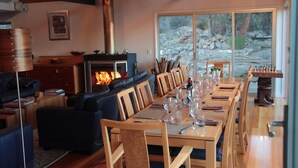 A dining area with a long wooden table set for six, surrounded by wooden chairs. A fireplace with a visible fire is in the background, and the room has a large window offering a view of the outdoors. - Blue Lake Lodge (Arthurs Lake)