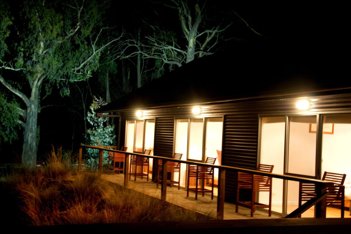 A wooden cabin with a balcony, furnished with chairs and tables, surrounded by trees.