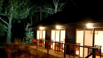 A wooden cabin with a balcony, furnished with chairs and tables, surrounded by trees.