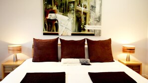 A neatly made bed with brown pillows and a white duvet, flanked by two bedside tables with lamps. Above the bed, there is a large abstract painting. - Blue Lake Lodge (Arthurs Lake)