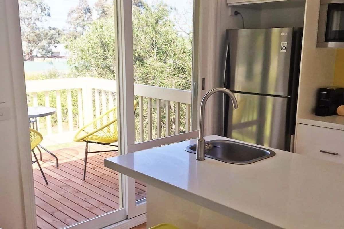 A kitchen with a stainless steel refrigerator, a sink, and a view of a wooden deck with chairs.