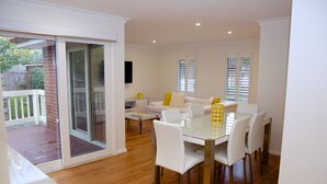 A modern dining area with a glass table and white chairs, a white sofa in the background, and a sliding glass door leading to an outdoor deck. - Yarra Glen Racecourse Apartments (Yarra Glen)