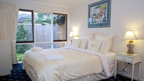 A neatly made bed with a white comforter and pillows, a bedside table with a lamp, and a window with curtains. - Yarra Glen Racecourse Apartments (Yarra Glen)
