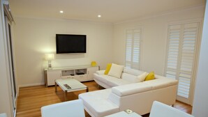 A modern living room with a white sofa, a glass coffee table, and a flat-screen TV mounted on the wall. - Yarra Glen Racecourse Apartments (Yarra Glen)