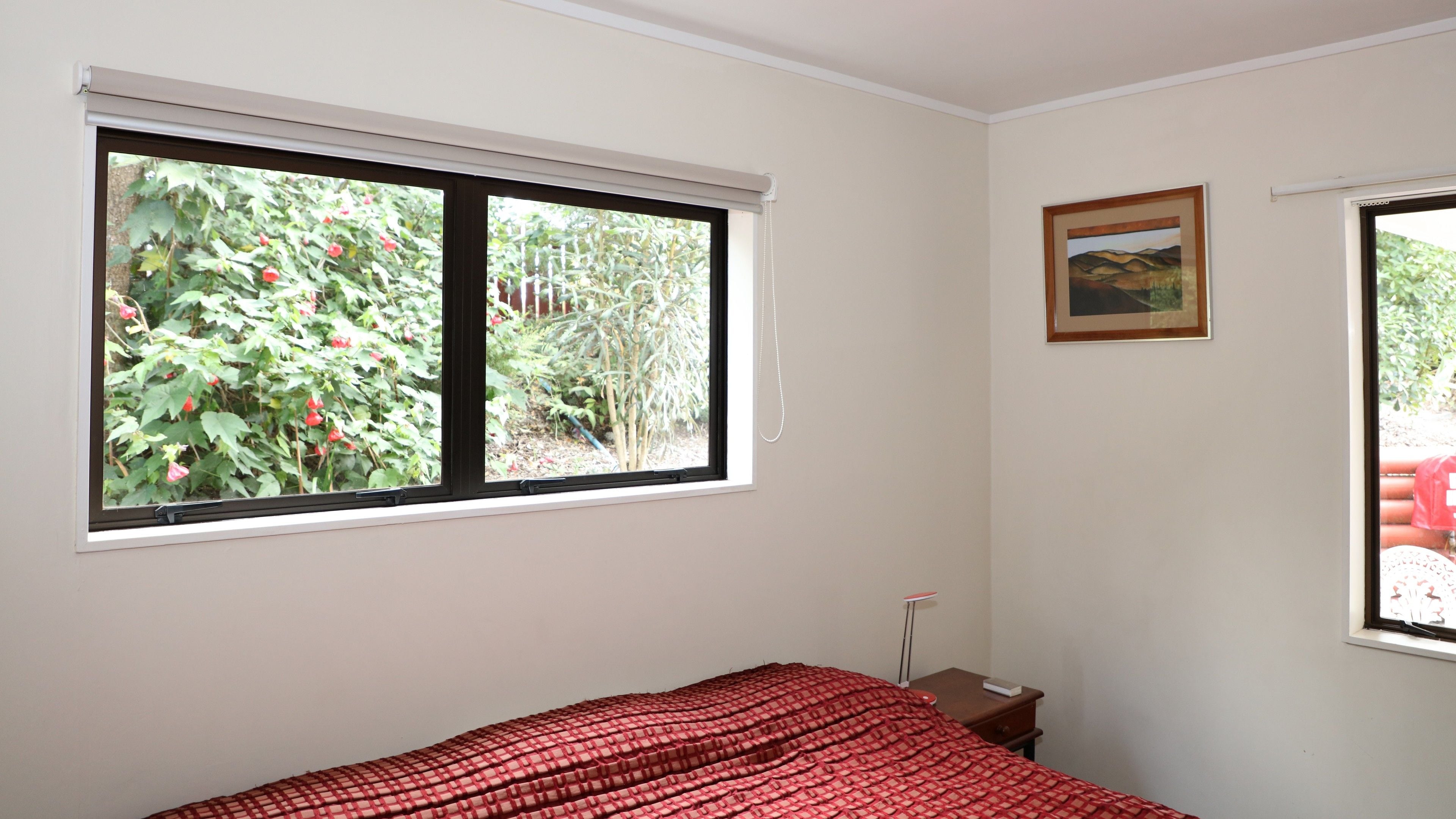 A bedroom with a large window, a bed with a red checkered bedspread, a small bedside table, and a framed picture on the wall.