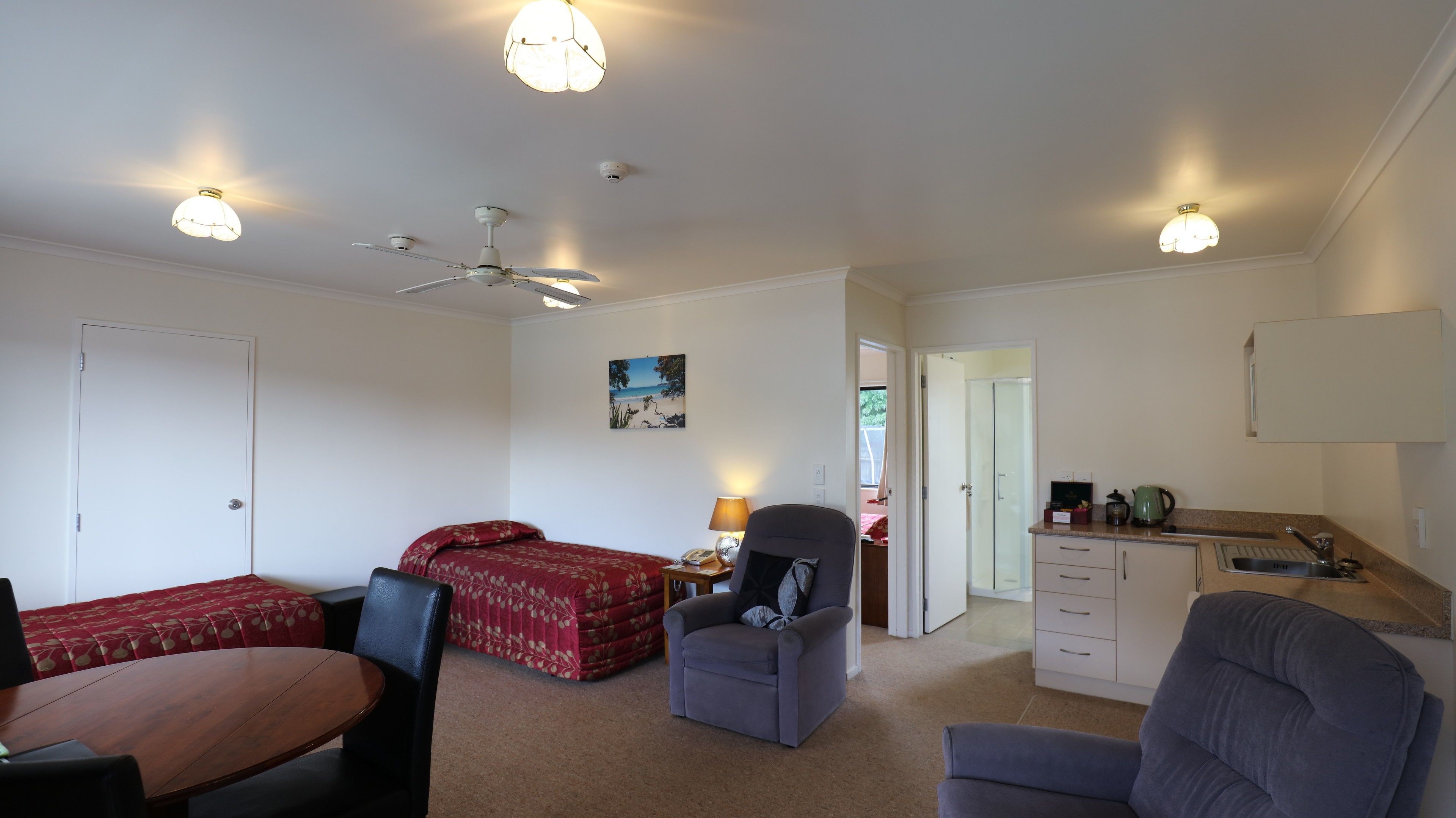 A compact room with a bed, two chairs, a desk, a ceiling fan, and a kitchenette.