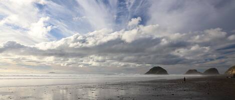 Une plage dotée d’un large rivage sablonneux, d’une eau calme et de formations rocheuses lointaines, le tout sous un ciel partiellement nuageux.