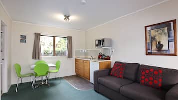 A compact living space with a kitchenette, a dining area with green chairs, a grey sofa with red patterned cushions, and a painting on the wall.