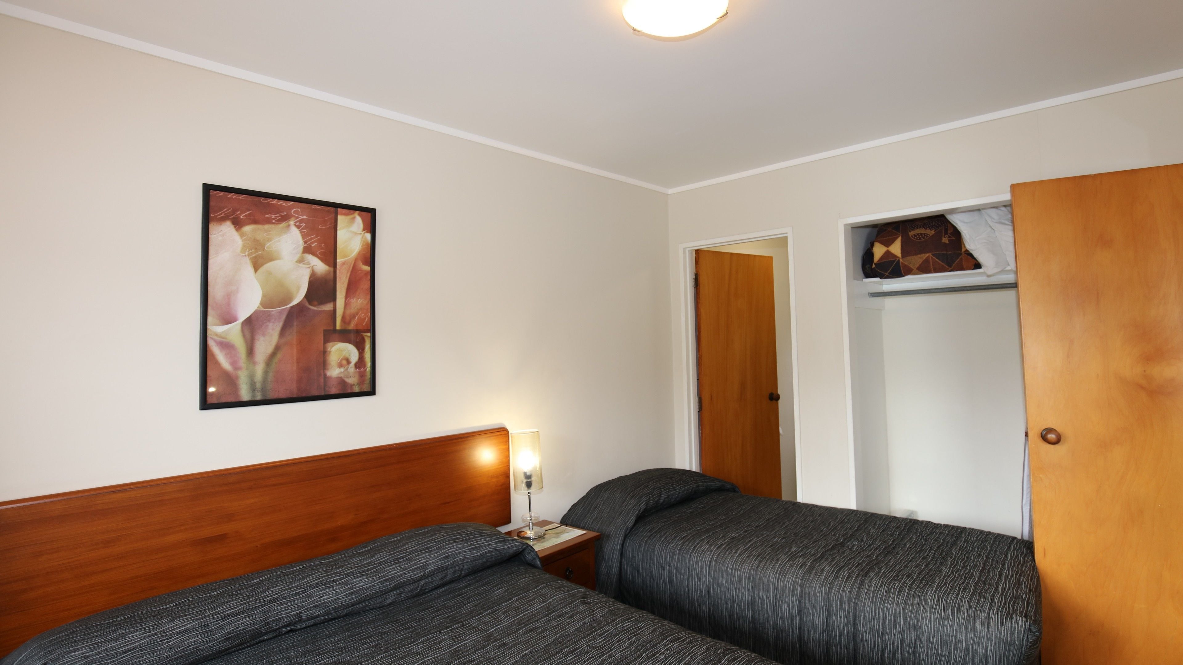 Habitación de hotel con dos camas, una mesita de noche con lámpara, un cabecero de madera, un cuadro floral enmarcado en la pared y un armario con un bolso estampado encima.