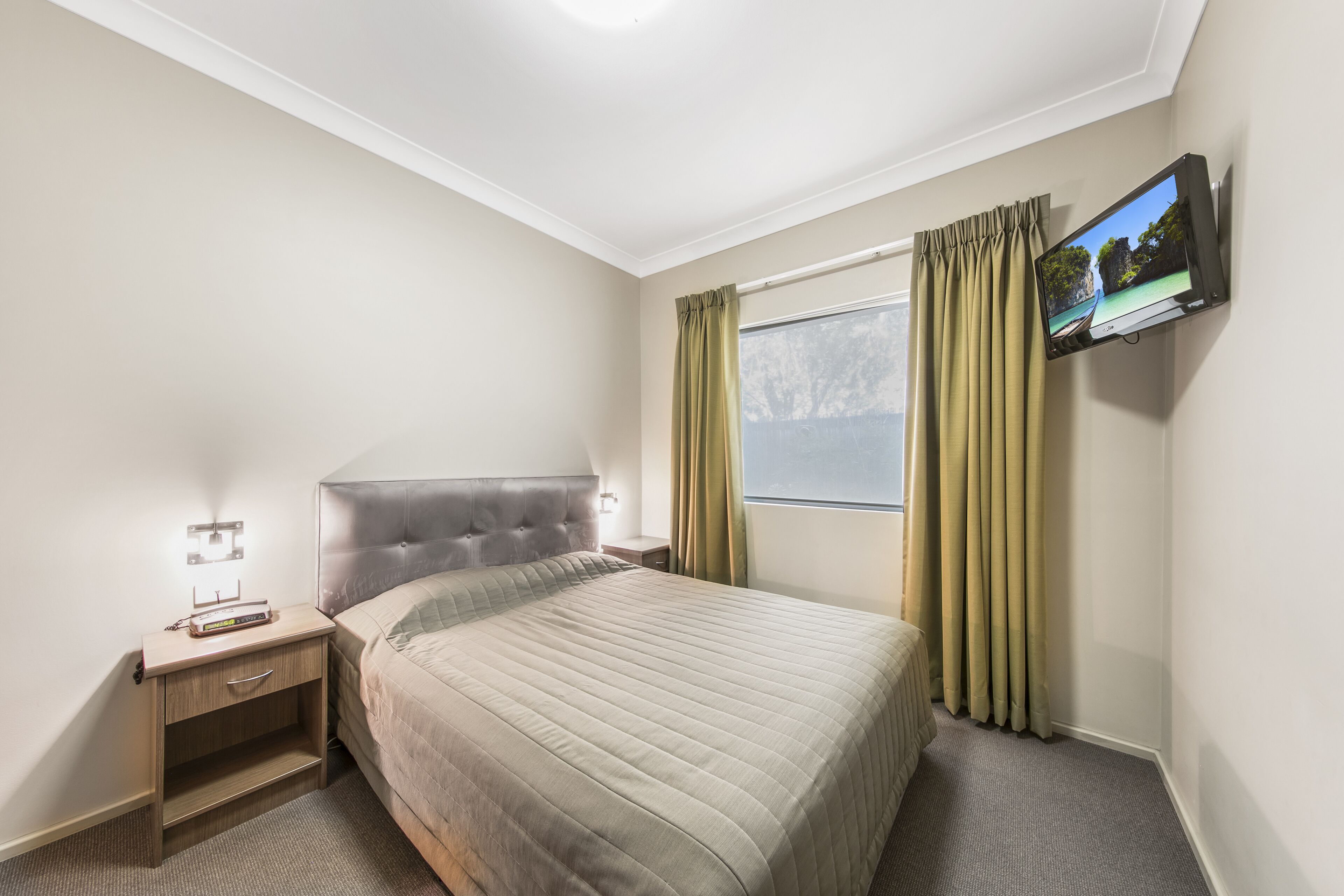 A hotel room with a bed, a nightstand, a TV mounted on the wall, and a window with curtains.