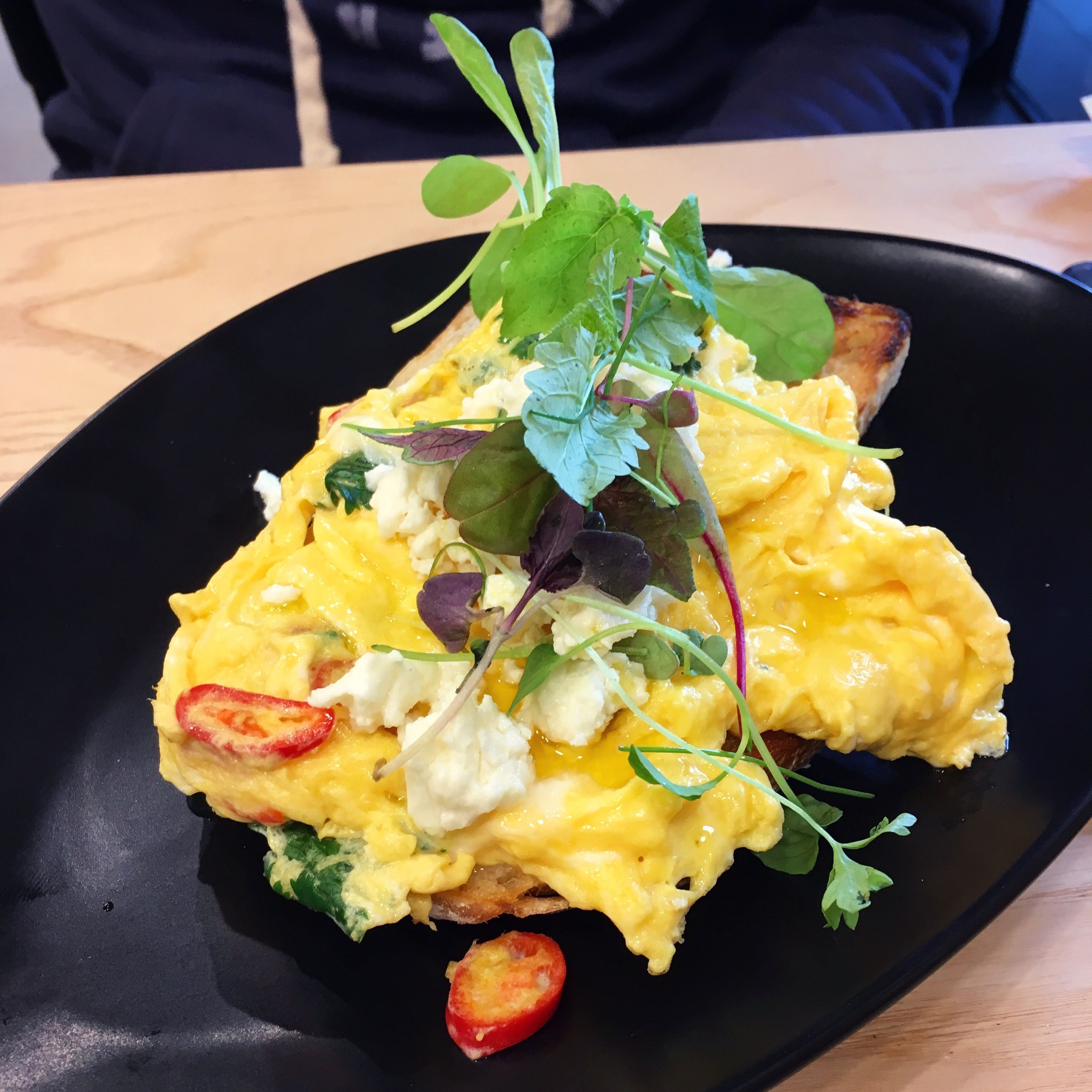 A black plate with a breakfast dish consisting of scrambled eggs, a slice of toast, and fresh greens.