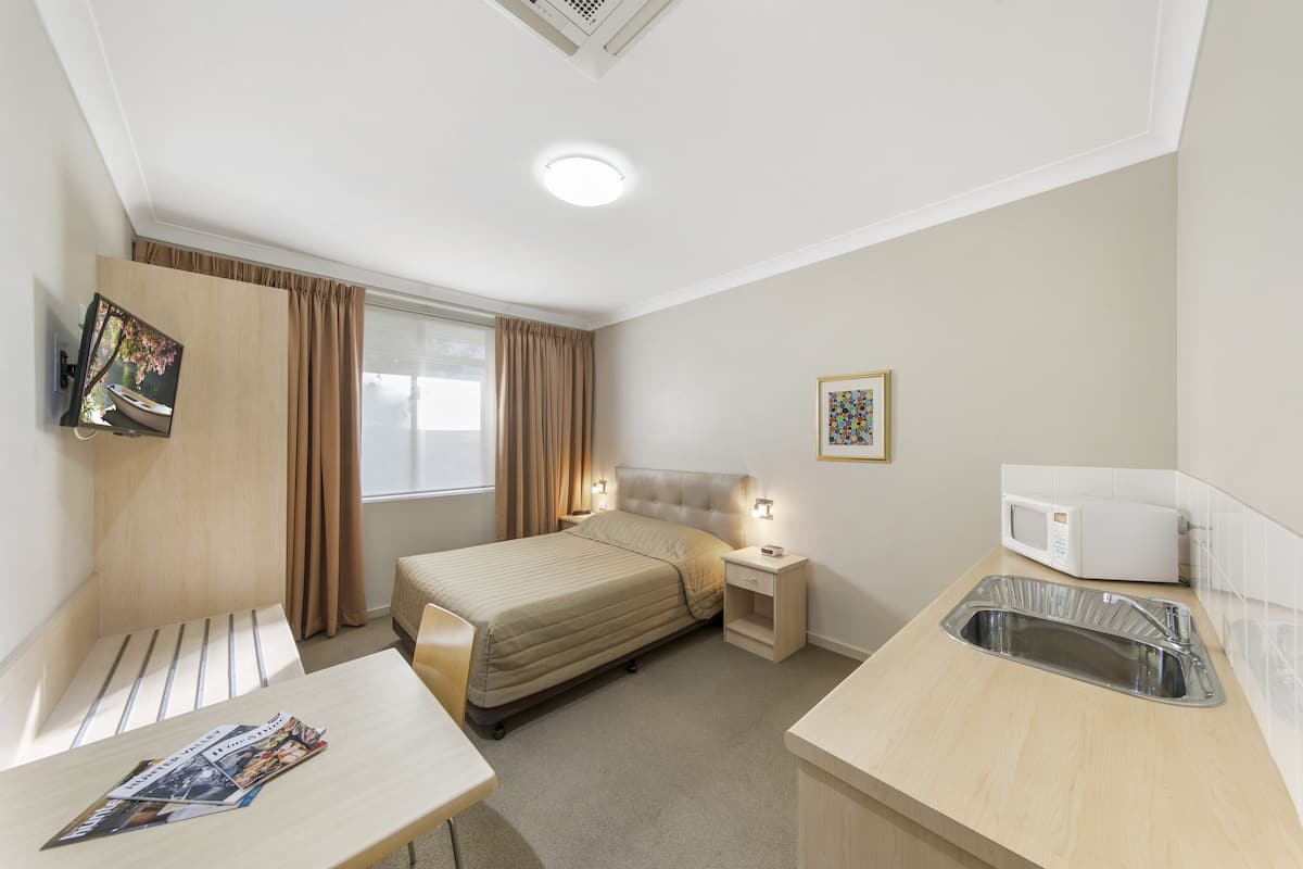 A hotel room with a bed, bedside table, lamp, TV, microwave, and a small sink.