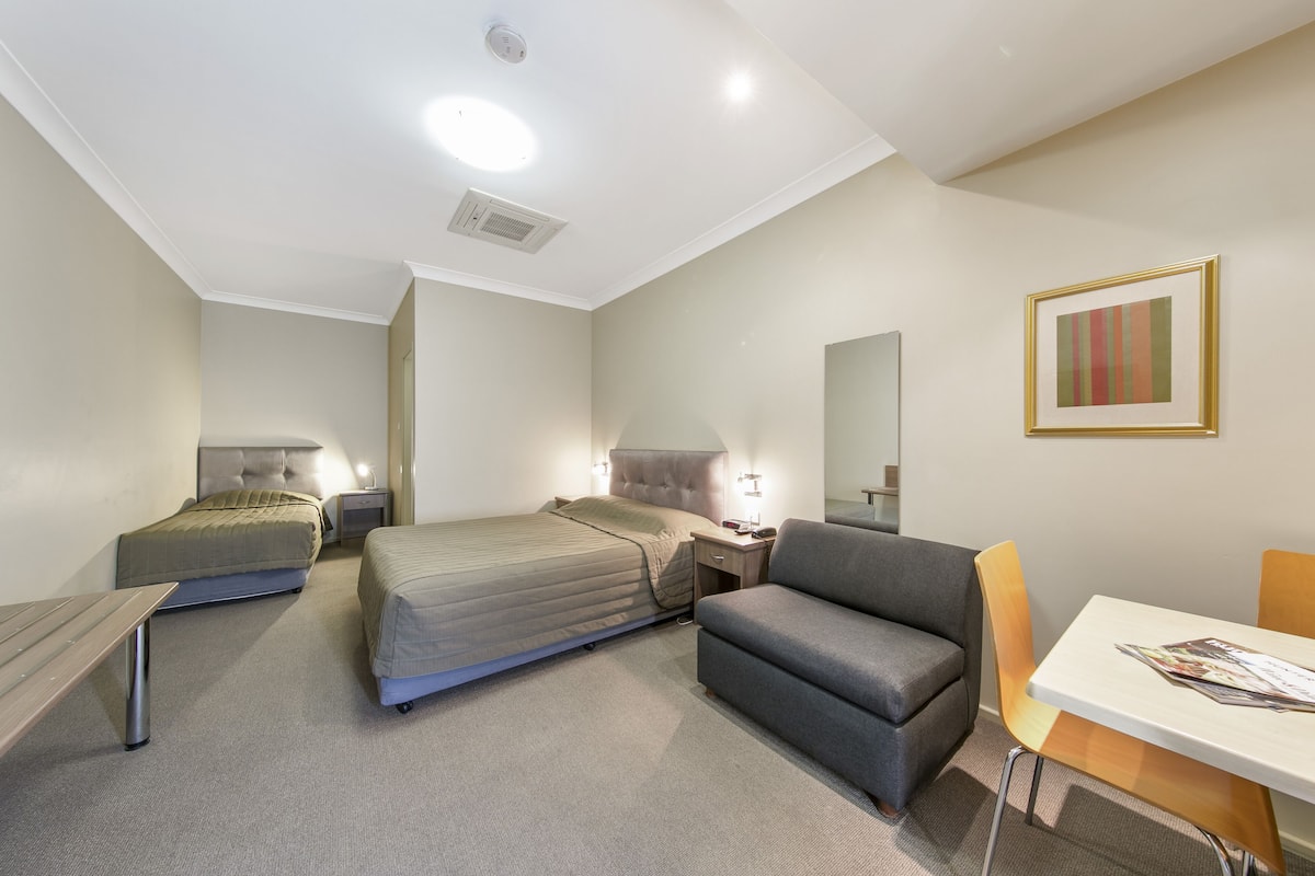 A hotel room with two beds, a desk, a chair, a table, a lamp, and a framed picture on the wall.