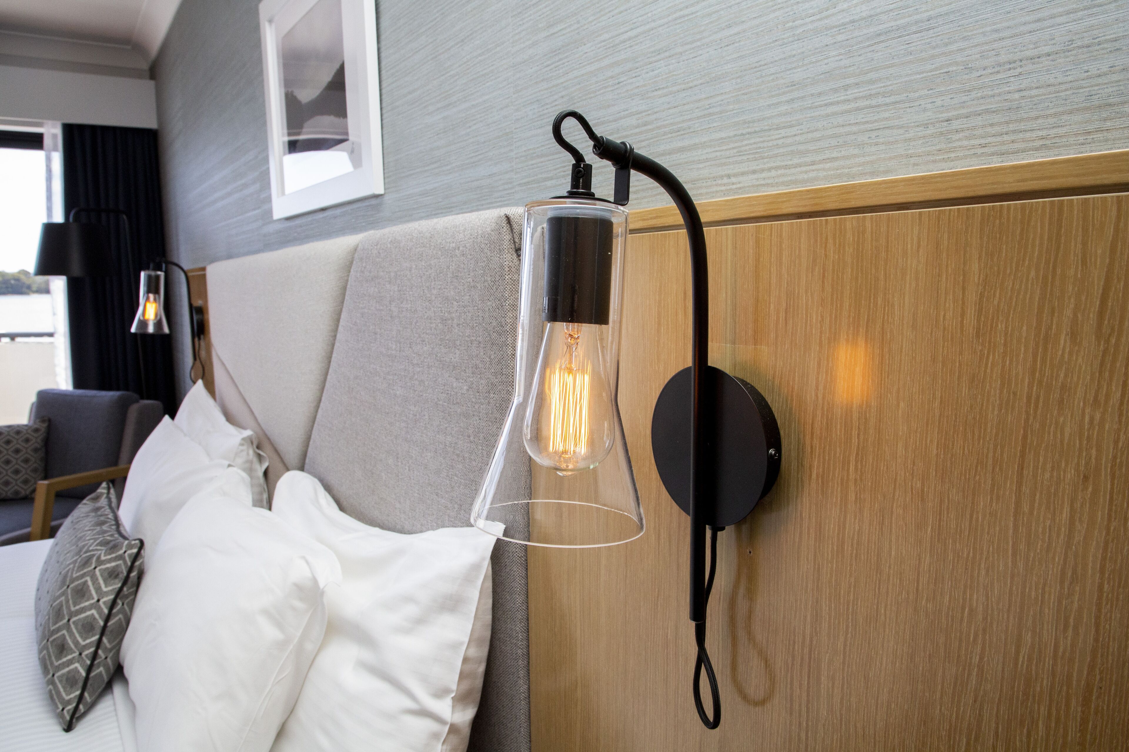 A modern hotel room with a clear glass pendant light fixture, a wooden headboard, and a view of the outdoors.
