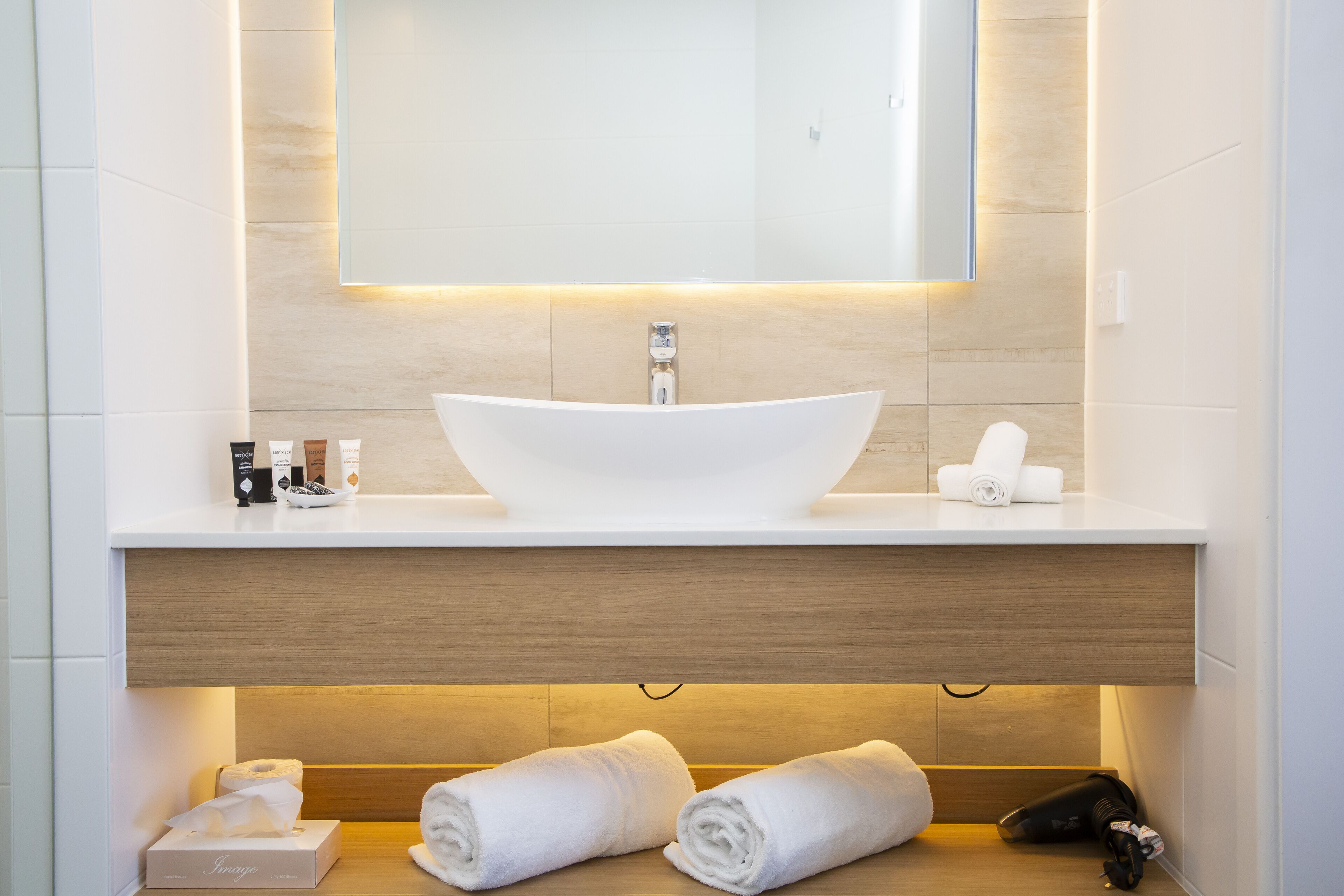 A modern bathroom with a large white sink, a mirror above it, and a wooden vanity below. There are two rolled towels on the floor and a box of tissues on the vanity.