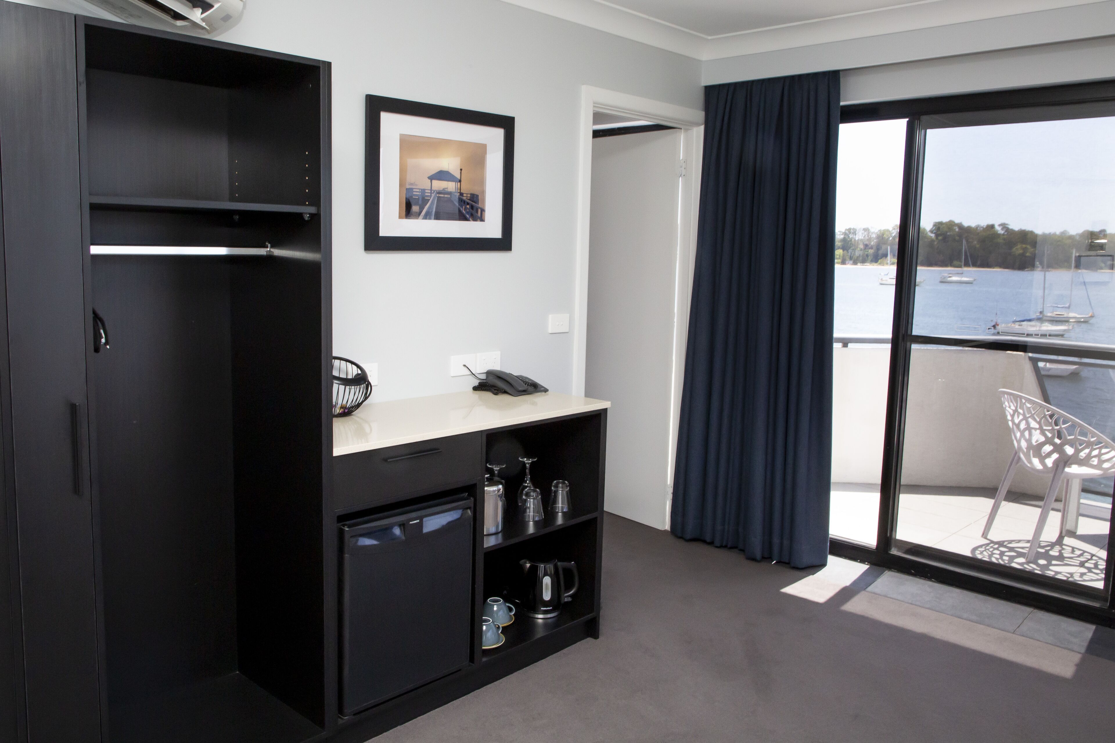 Ein modernes Hotelzimmer mit Balkon, einem gerahmten Bild an der Wand und einem Schreibtisch mit Telefon und Wasserkocher.