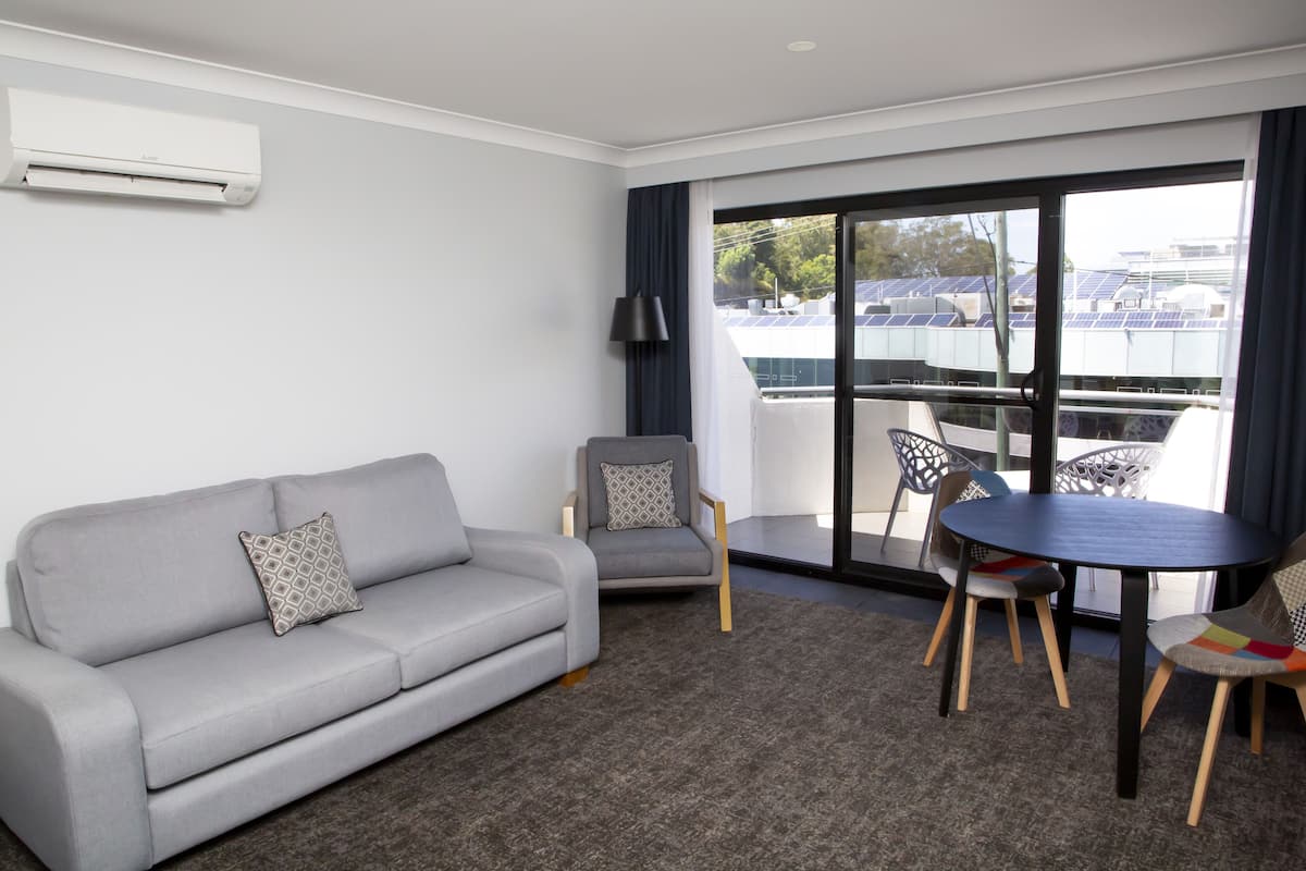 A modern hotel room with a grey sofa, a small table, and two chairs. There is a large window with a view of solar panels outside.