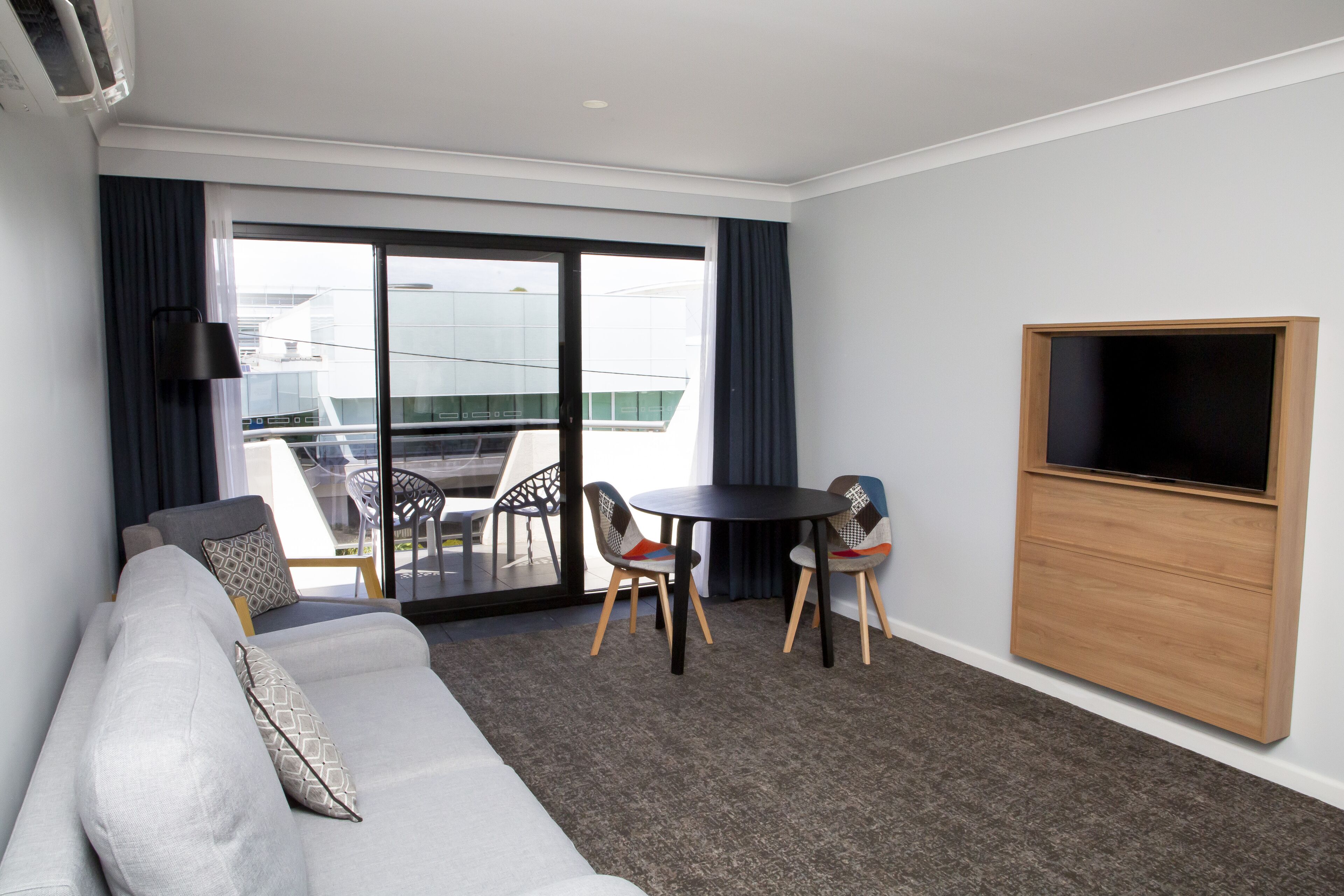 A modern hotel room with a grey sofa, two chairs, a small round table, a TV, and a balcony with a glass door.