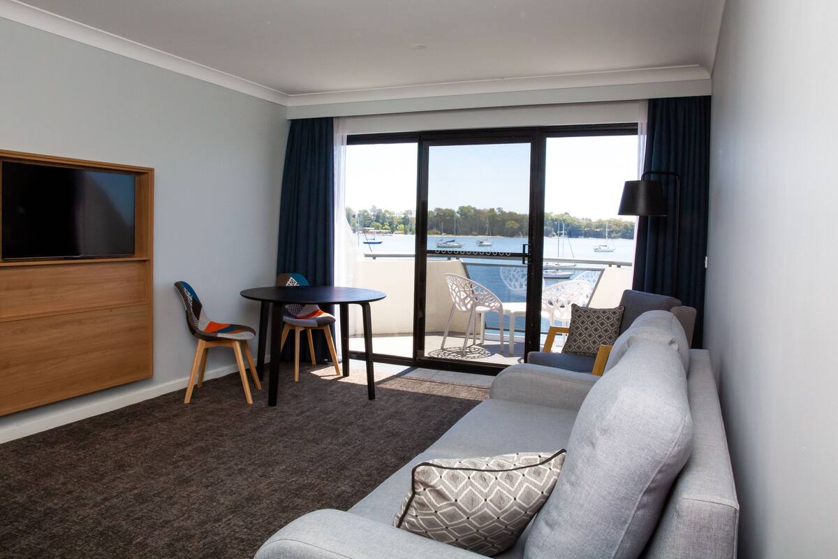 A hotel room with a sofa, a TV, a small table with chairs, and a balcony with a view of the water.