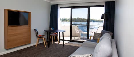 A hotel room with a sofa, a TV, a small table with chairs, and a balcony with a view of the water.